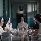 Mönchinnen sitzen um einen langen Tisch. Eine hält in der Mitte die Hände gefaltet. Oberlichter werfen Schatten.