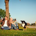 Ein Verkehrsschild zeigt "Gerolstein 10 km, Neroth 3 km". Zwei Männer sitzen trinkend im Gras, ein Hund läuft vorbei.
