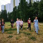 Mehrere junge Männer in lässiger Kleidung gehen über eine Wiese, im Hintergrund stehen hohe Wohngebäude und Bäume.