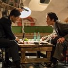 Zwei Männer in Anzügen sitzen angespannt an einem niedrigen Tisch mit Getränken, umgeben von einer intensiven Atmosphäre.