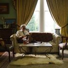 Ein älterer Mann sitzt lesend auf einem Sofa in einem eleganten Raum mit Vorhängen, antiken Möbeln und Gemälden an der Wand.