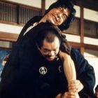 Zwei Männer in einem intensiven Kampf in einem traditionellen japanischen Raum. Einer trägt ein dunkles Gewand mit einem runden Symbol.