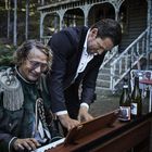 Zwei Männer an einem Klavier, einer lächelnd in historischem Kostüm, der andere im Anzug. Im Hintergrund ein altes Gebäude.