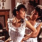 Zwei Frauen in viktorianischen Kleidern; eine hilft der anderen beim Schnüren des Korsetts in einem elegant eingerichteten Raum.