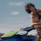 Ein Paar auf einem Jetski; die Frau macht ein Selfie mit dem Smartphone, während sie im Bikini hinter dem Mann sitzt.