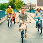 Drei Männer fahren auf Fahrrädern über eine Brücke in einer Vorstadt. Ein Fahrrad hat einen Korb mit einem Tuch darin.