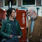 Text: "13-4271" auf rotem Fahrzeug, darüber drei Personen im Gespräch. Gebäude im Hintergrund.