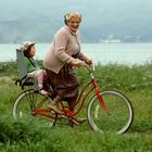 Eine ältere Person fährt ein rotes Fahrrad mit einem Kind im Kindersitz durch eine grüne Wiese, im Hintergrund ist Wasser.