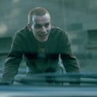 Ein Mann mit kurzem Haar lehnt sich über eine Oberfläche und lächelt in einer urbanen Umgebung.