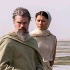 Ein Mann mit grauem Bart und eine Frau mit Kopftuch stehen in einer Wüstenlandschaft. Sie tragen schlichte Gewänder.