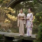 Zwei Frauen in Kimonos stehen auf einer Brücke in einem traditionellen japanischen Garten mit üppigem Grün und einem Teich.