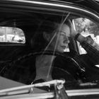 Eine Frau sitzt nachdenklich am Steuer eines alten Autos, mit der Hand am Kopf, schwarz-weiß.