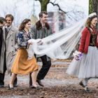 Eine Gruppe lächelnder Menschen in Vintage-Kleidung, begleitet ein Brautpaar im Freien. Die Braut trägt einen Schleier.