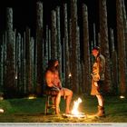 Zwei Männer in einem Wald aus massiven Holzpfählen, eine Flamme zwischen ihnen, Totenschädel auf der linken Seite.