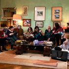 Eine Gruppe von Menschen lächelt und öffnet Geschenke in einem gemütlichen Wohnzimmer mit Weihnachtsbaum.