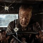 Ein bärtiger Mann, dreckig und entschlossen, fährt ein Auto mit zerschossener Windschutzscheibe.