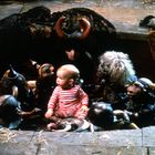 Ein Baby in rot-weiß gestreifter Kleidung sitzt umgeben von kleinen Kreaturen mit Hörnern und Fell auf einem Boden.