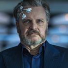 Ein Mann mit grauem Haar und Bart trägt ein Gerät mit Elektroden an der Stirn, in formeller Kleidung. Im Hintergrund unscharfe Lichter.