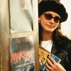 Eine Person in Lederjacke und Baskenmütze hält Bücher mit Text auf dem Cover in der Hand. Sie trägt eine Sonnenbrille.