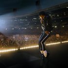 Ein Künstler tritt auf einer Bühne auf. Ein Publikum mit Schildern jubelt. Scheinwerfer strahlen in die Menge.