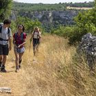 Drei Personen wandern auf einem Pfad durch eine trockene, grasbewachsene Landschaft, umgeben von Bäumen und Felsen.