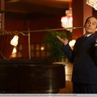 Person in elegantem Anzug steht neben einem Klavier, hebt die Hände fragend, während ein jüngerer Mann am Klavier sitzt.