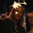 Zwei Menschen halten Wunderkerzen in der Dunkelheit. Links ist eine Frau, rechts ein Kind mit Brille. Beide lächeln.