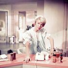 Keine sichtbaren Texte. Eine blonde Frau in einem hellen, eleganten Raum; neben ihr ein Telefon und Flaschen auf einer Theke.