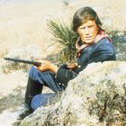 Ein Mann in Uniform mit Gewehr kauert hinter einem Felsen in einer trockenen Landschaft.