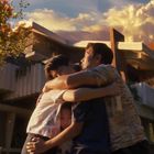 Eine Familie umarmt sich innig vor einem modernen Haus bei Sonnenuntergang, umgeben von Bäumen und warmem Licht.