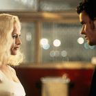 Eine blonde Frau in einem weißen Top sieht einem Mann mit dunklem Haar gegenüber. Der Hintergrund ist verschwommen.