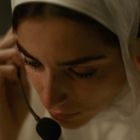 Eine Frau trägt ein Headset, hält es mit beiden Händen und wirkt konzentriert. Sie trägt ein weißes Kopftuch.