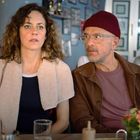 Zwei Personen, eine Frau und ein Mann mit Brille und Mütze, sitzen nebeneinander in einem Café mit Getränken auf dem Tisch.