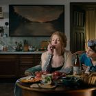 Eine gemütliche Küche mit zwei Frauen am Tisch, gedeckt mit Essen. Im Hintergrund ein Landschaftsgemälde.