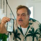 Ein Mann im Hemd mit Palmenmuster hält ein Smartphone nahe ans Gesicht. Im Hintergrund eine Treppe und Pflanzen.