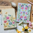 Zwei Notizbücher mit bunten Blumenstickereien und Schmetterlingen, auf Holzuntergrund und neben Blumenvasen liegend.