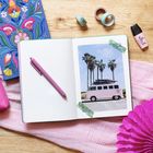 "Be You Tiful" steht auf einem rosa Fläschchen. Ein Notizbuch mit einem Bild von Palmen und einem pinken Bus ist zu sehen.
