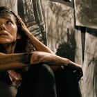 Verschmutzte Frau sitzt erschöpft am Boden, umgeben von düsteren Zeichnungen an der Wand, schaut nach oben.