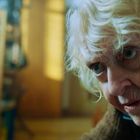 Eine ältere Person mit lockigem, grauem Haar und intensivem Blick in Nahaufnahme. Verschwommener Hintergrund mit Möbeln.