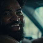 Eine Person sitzt lächelnd im Auto und schaut zur Seite. Die Szene ist sonnig und leicht verschwommen.