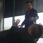 Keine sichtbaren Texte. Zwei Männer in einem modernen Büro, einer liegt auf dem Tisch, ein anderer steht bedrohlich über ihm. Ein Dritter ist im Hintergrund am Handy.