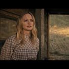 Keine Texte vorhanden. Eine Frau mit langen blonden Haaren steht vor einer Holzwand und blickt nachdenklich.