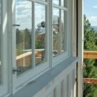 Eine Frau mit Fernglas steht auf einem Balkon, umgeben von Bäumen, Fenster spiegelt die Szene.