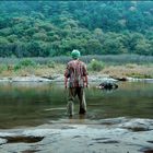 Zu sehen ist eine Person mit buntem Hemd und grünem Haar, die in einem flachen Fluss steht, umgeben von Wald.