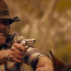 Ein Mann in kariertem Hemd und Cowboyhut richtet eine Pistole. Er hat einen angespannten Gesichtsausdruck.