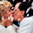 Ein Paar in Hochzeitskleidung küsst sich herzlich, umgeben von herabfallenden Blütenblättern.