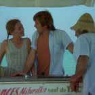 „Nos Glaces Naturelles“ steht auf einem Wagen. Drei Personen unterhalten sich fröhlich am Strand.