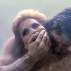 Ein Mann hält einer Frau von hinten den Mund zu, während Wasser über sie fließt. Die Szene wirkt intensiv und emotional.