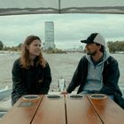 Zwei Menschen sitzen auf einem Boot vor einem Tisch. Im Hintergrund ist ein Fluss und eine urbane Landschaft sichtbar.