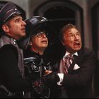 Drei Männer schauen überrascht oder ängstlich. Einer trägt eine Uniform, der mittlere einen schwarzen Helm.